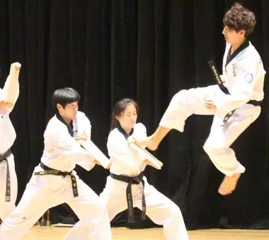 Students performing a karate demonstration on stage, with one martial artist executing a flying kick against a black backdrop.
